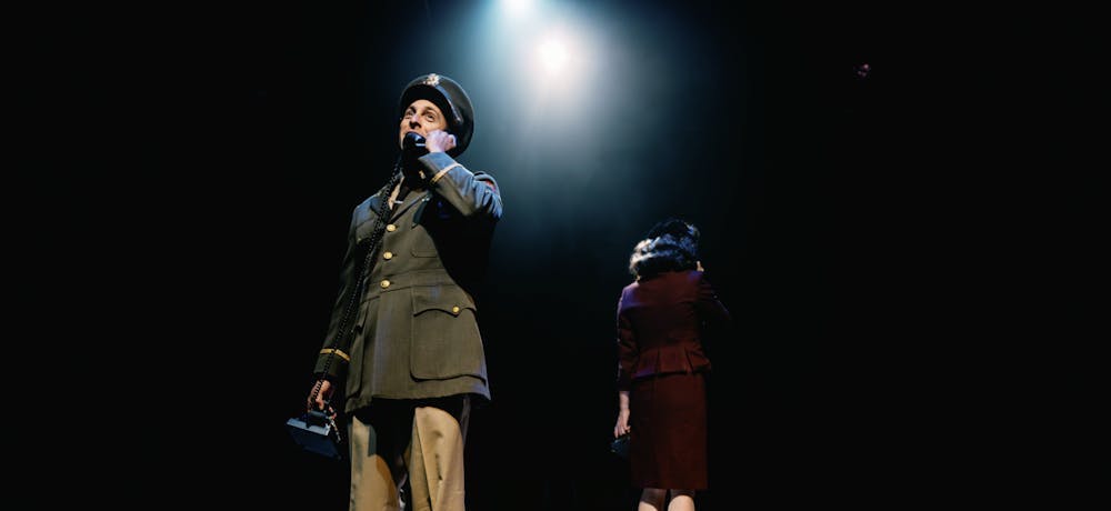 A man in military uniform faces the camera and holds a vintage phone to his ear; a woman wearing maroon stands behind him with her back facing the camera.