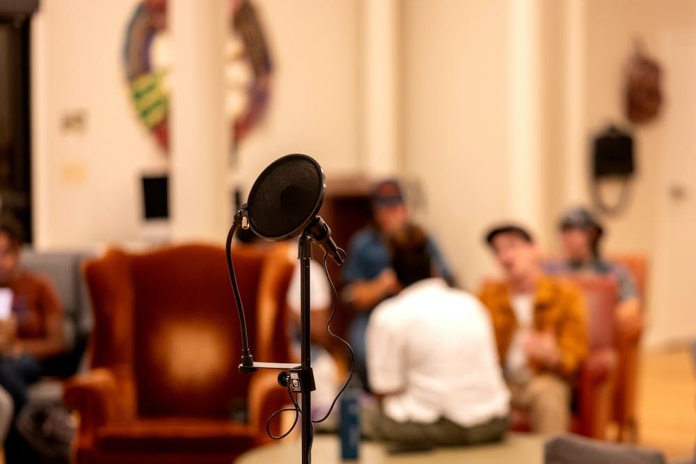 A microphone stands against a blurry background, which includes furniture and a handful of sitting people.
