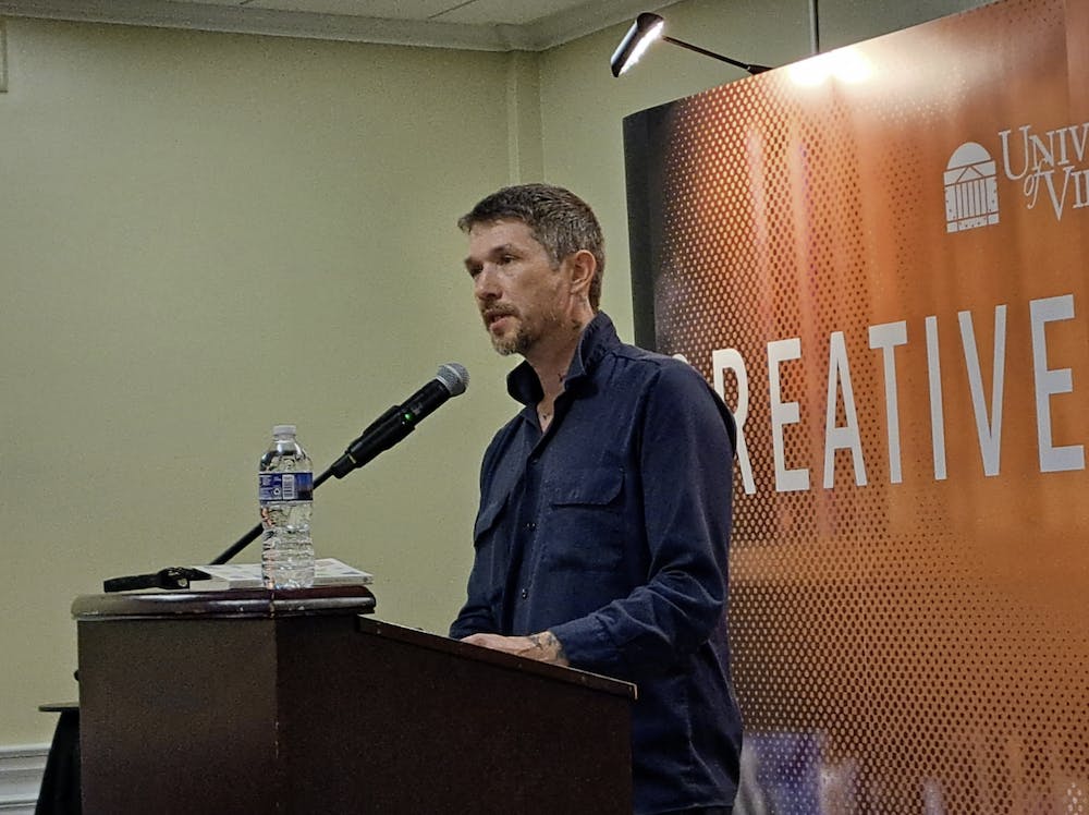 Jesse Ball, wearing a blue long sleeve shirt, stands at a lectern and speaks into a microphone. 