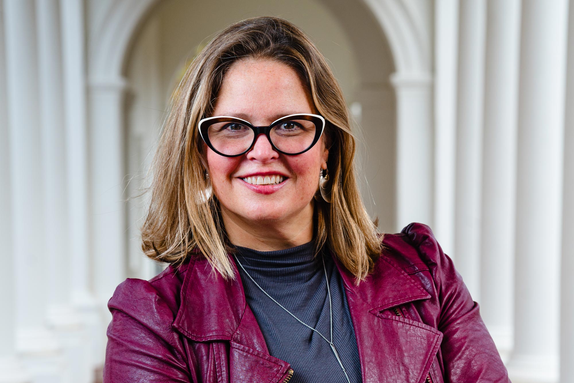 Karen Elizabeth Milbourne smiles at the camera wearing glasses, a red jacket, and a gray turtleneck. 