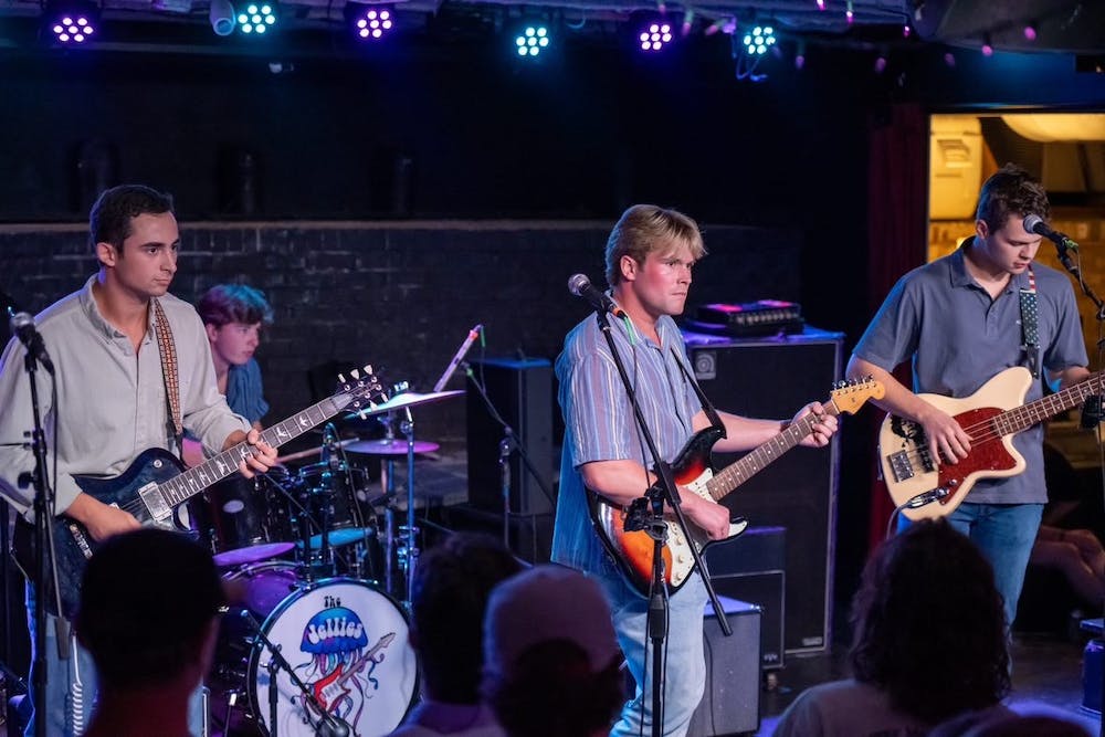 Three men holding guitars stand on a stage in front of another playing the drums. The drum set features a drawing of a jellyfish and the words "The Jellies."