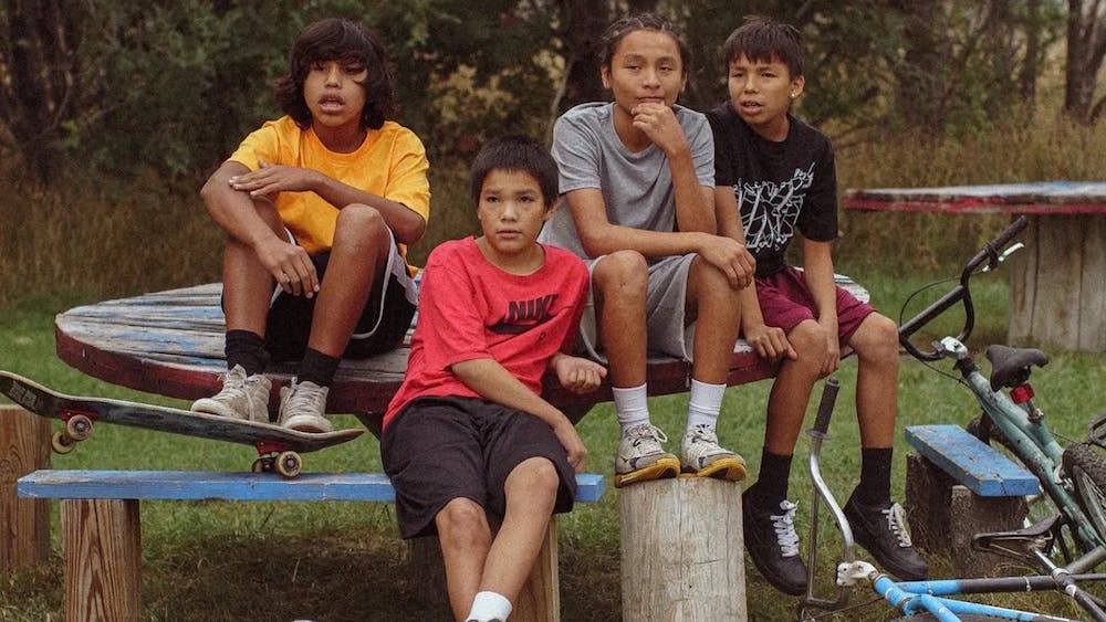 Four children in casual clothes sit on a wooden table outdoors. 