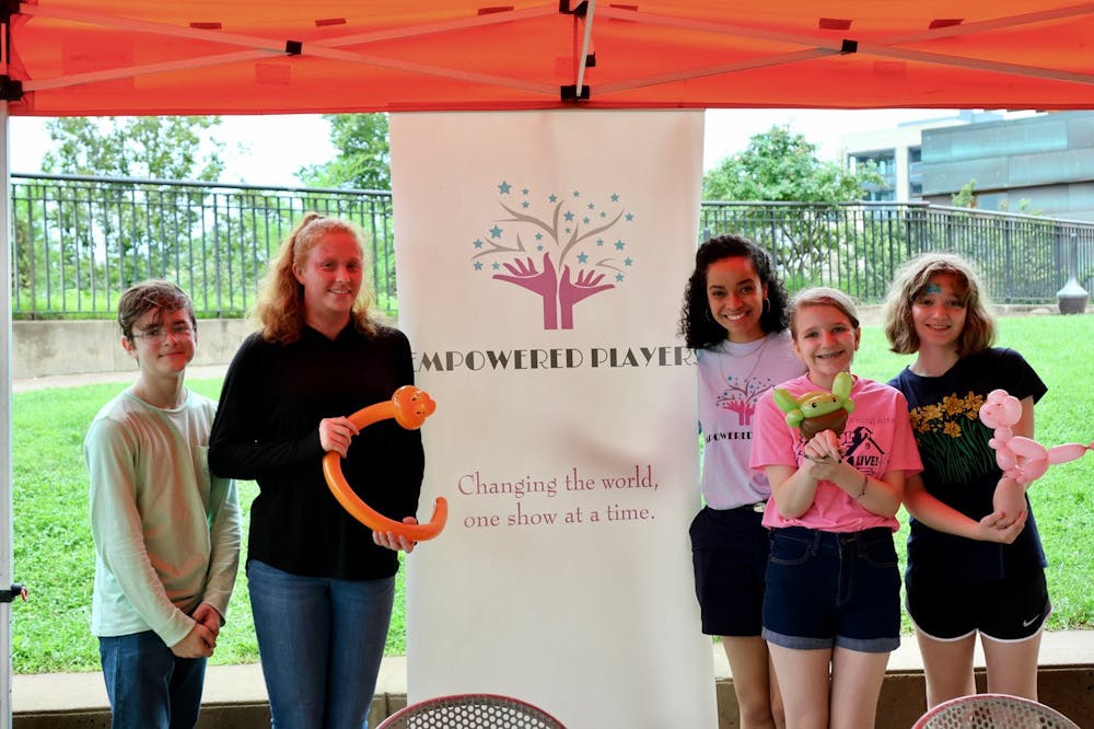 Jessica Harris smiles and stands next to a group of students in front of a banner that reads "Empowered Players" and "Changing the world, one show at a time." Several of the students hold balloon animals.