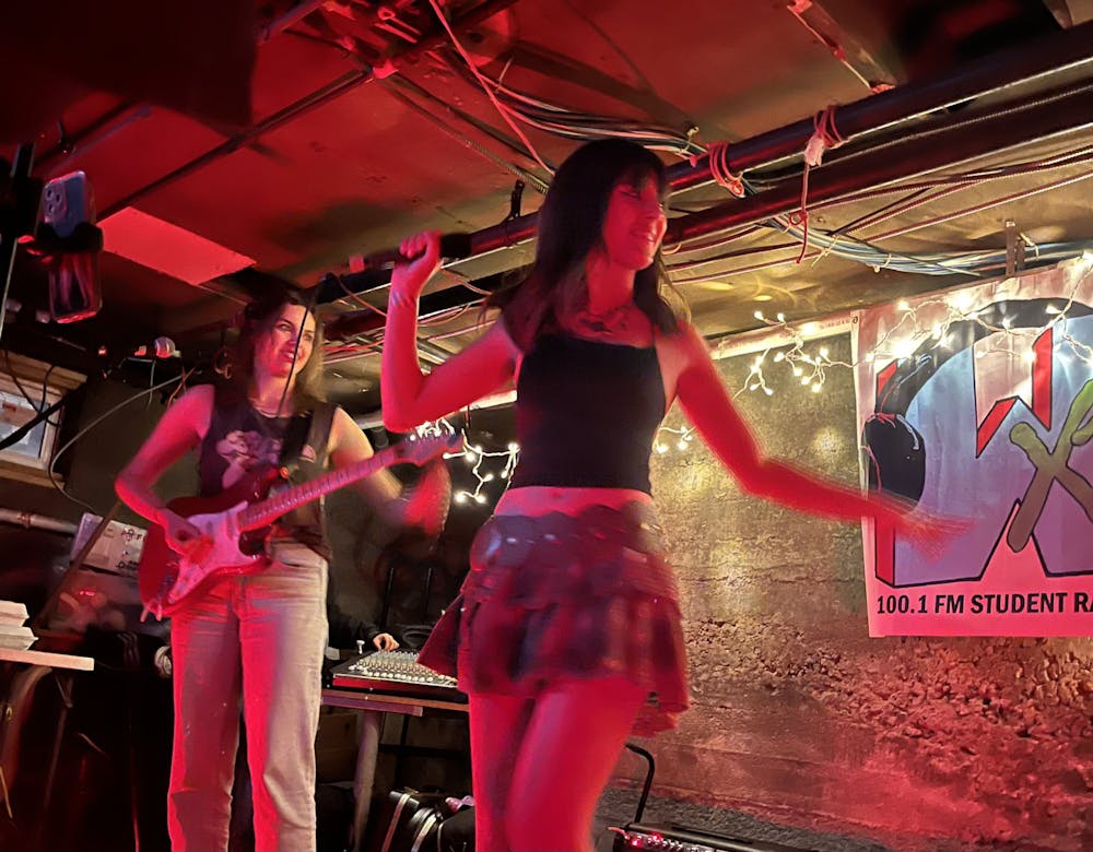 Two members of Fair Verona perform in a basement lit by string lights — one plays an electric guitar while the other holds a microphone.