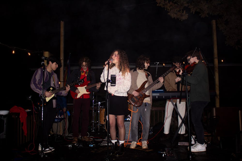  Six musicians play on a stage — two play guitars, one plays a bass, one plays a keyboard, one plays a fiddle, and one sings into a microphone.