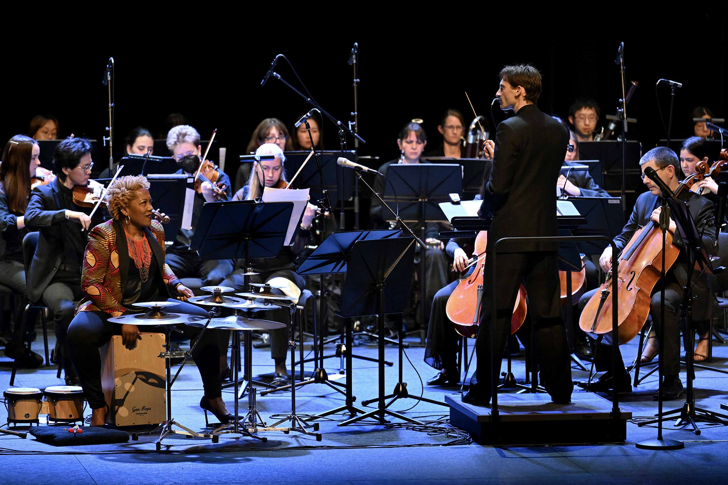 A group of musicians of the Charlottesville Symphony sit onstage under a blue light, 