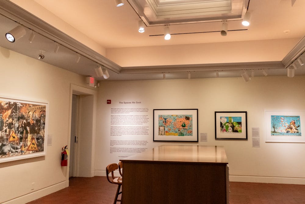 A photo of the Fralin's exhibit titled "The Spaces We Seek"; four pieces are shown on the walls next to a long passage on the wall detailing the aims of the exhibit (not legible in photo). A table sits in the middle of the room with tall chairs next to it.