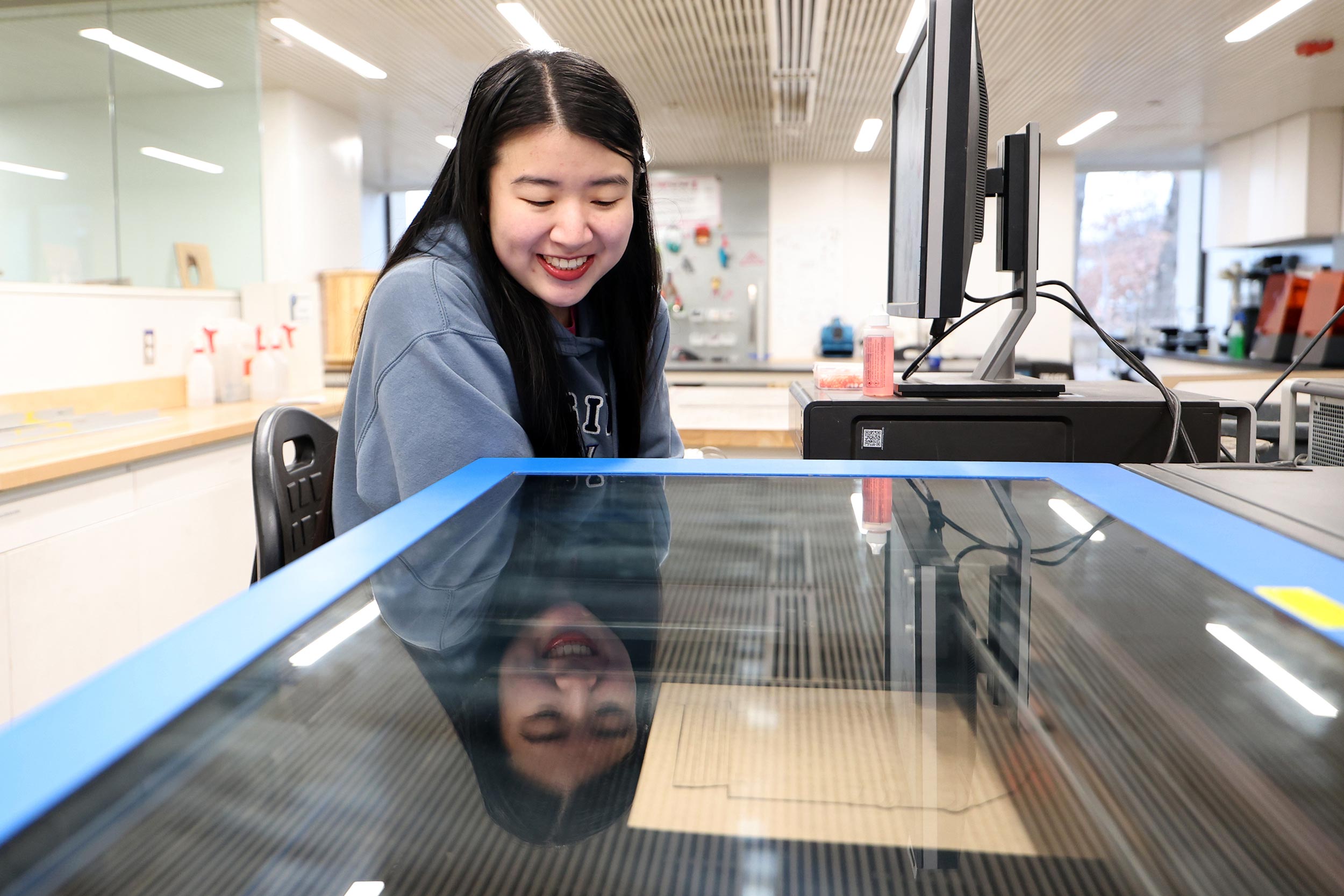 Timmins sits in front of a computer and looks down at a machine, smiling.