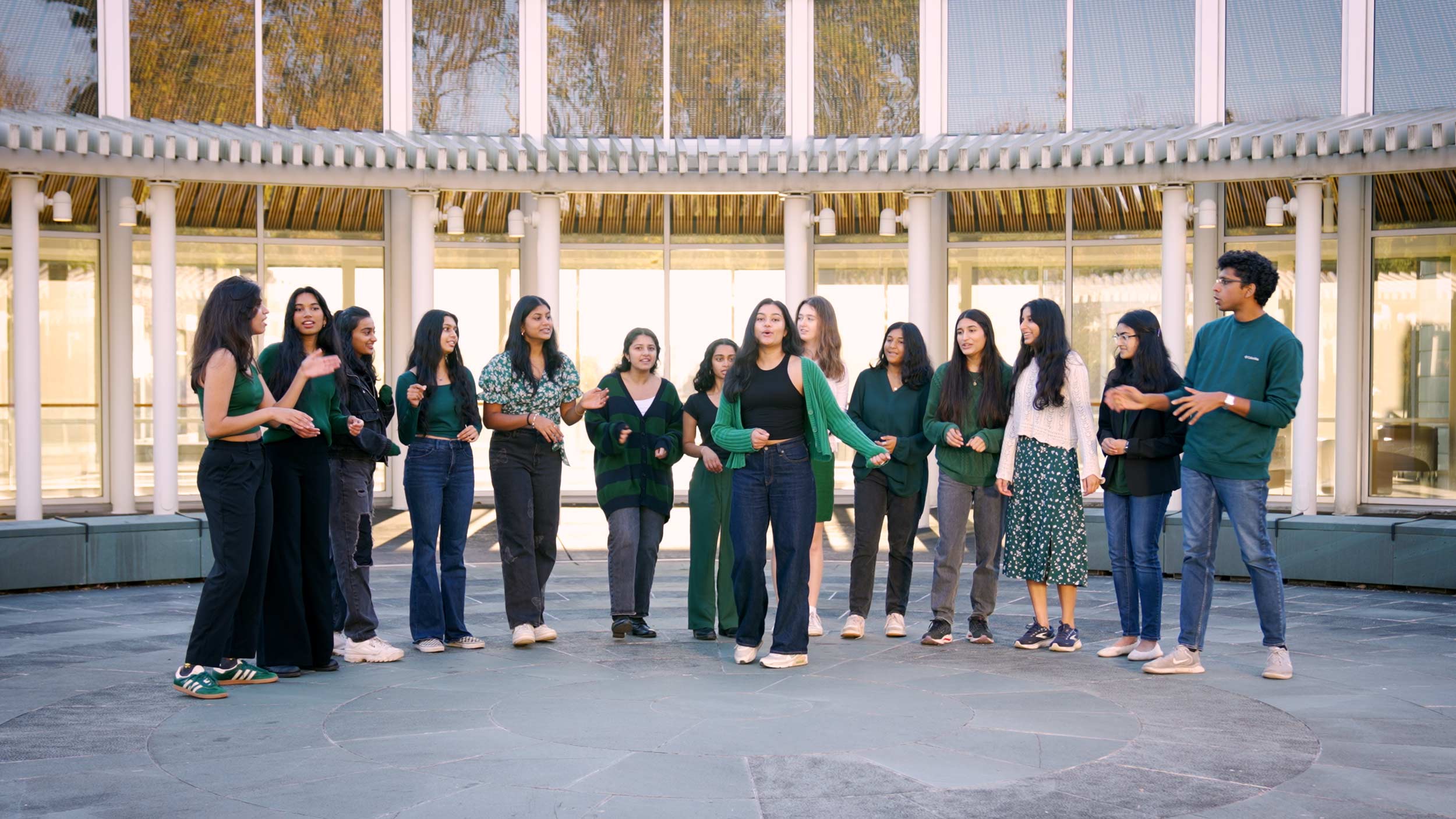 The members of Ektaal A Cappella, fourteen people wearing green, stand in an arc formation with a soloist in the middle outside of Nau Hall.