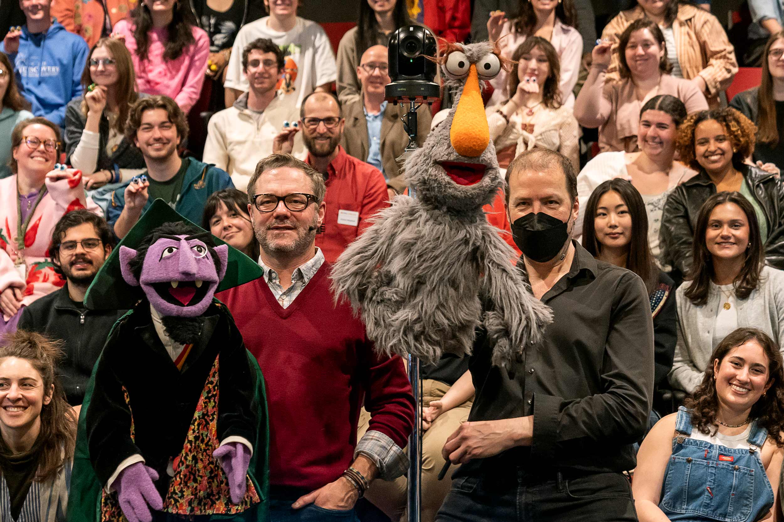 Matt Vogel and Jason Weber each hold a muppet while posing in front of a smiling audience.