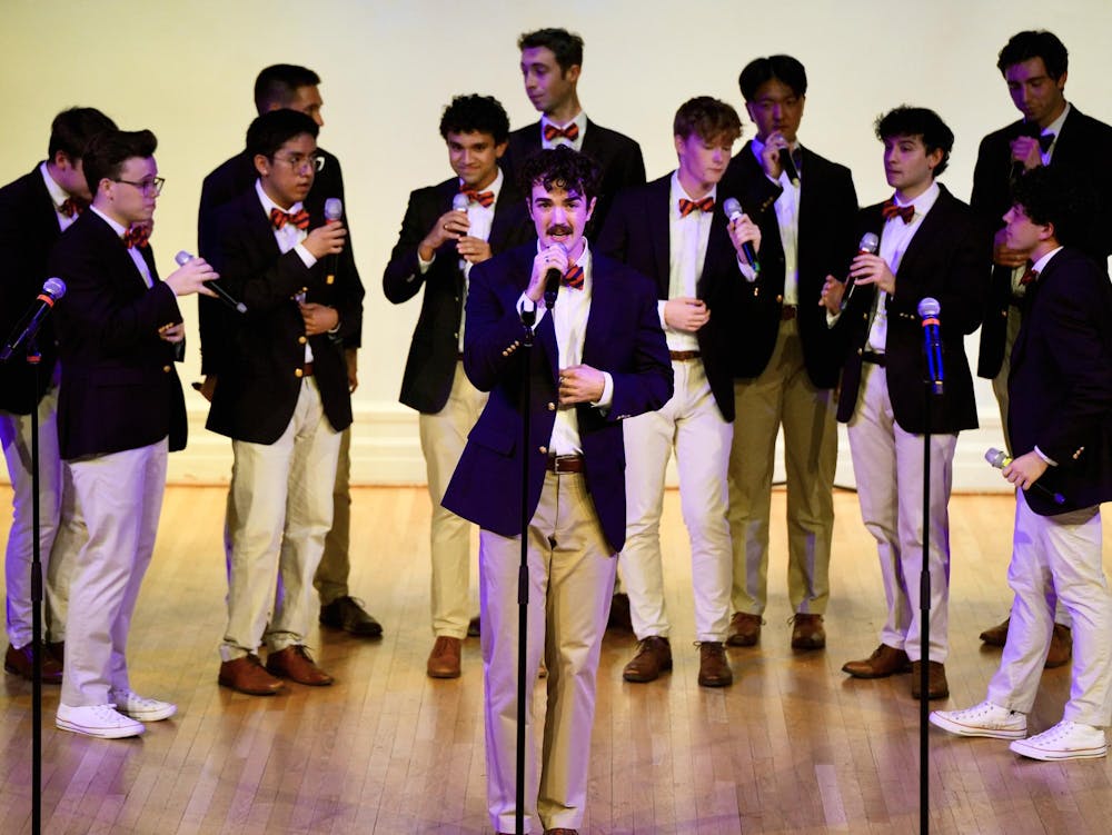 The members of the Virginia Gentlemen stand onstage in an arc formation with one soloist in the center. Each member wears a suit with a blue-and-orange striped bowtie.