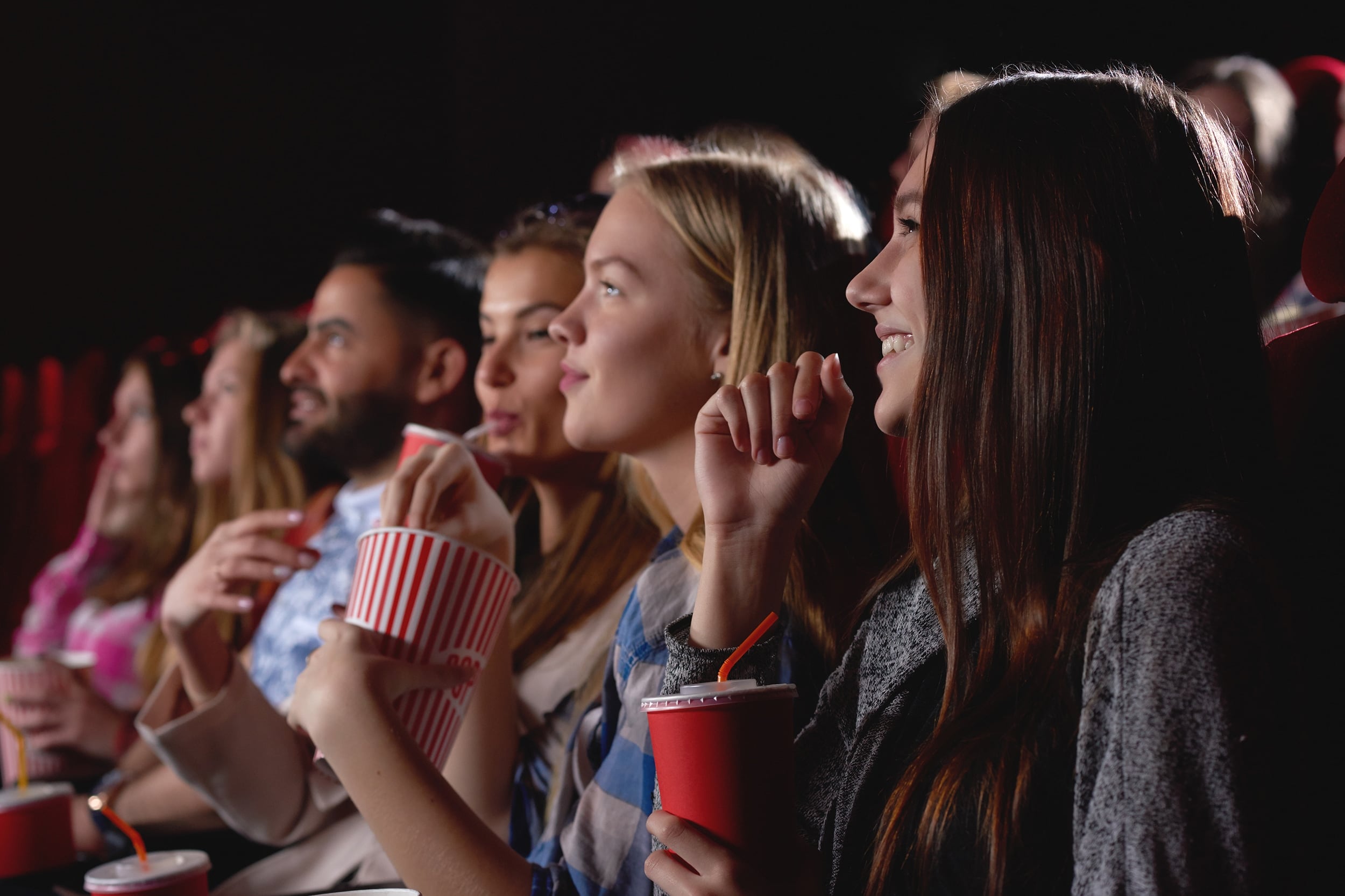 A row of people sit in a movie theater, watching a film: one reaches into a bucket of popcorn, while another sips a drink. The one closest to the camera smiles up at the screen.