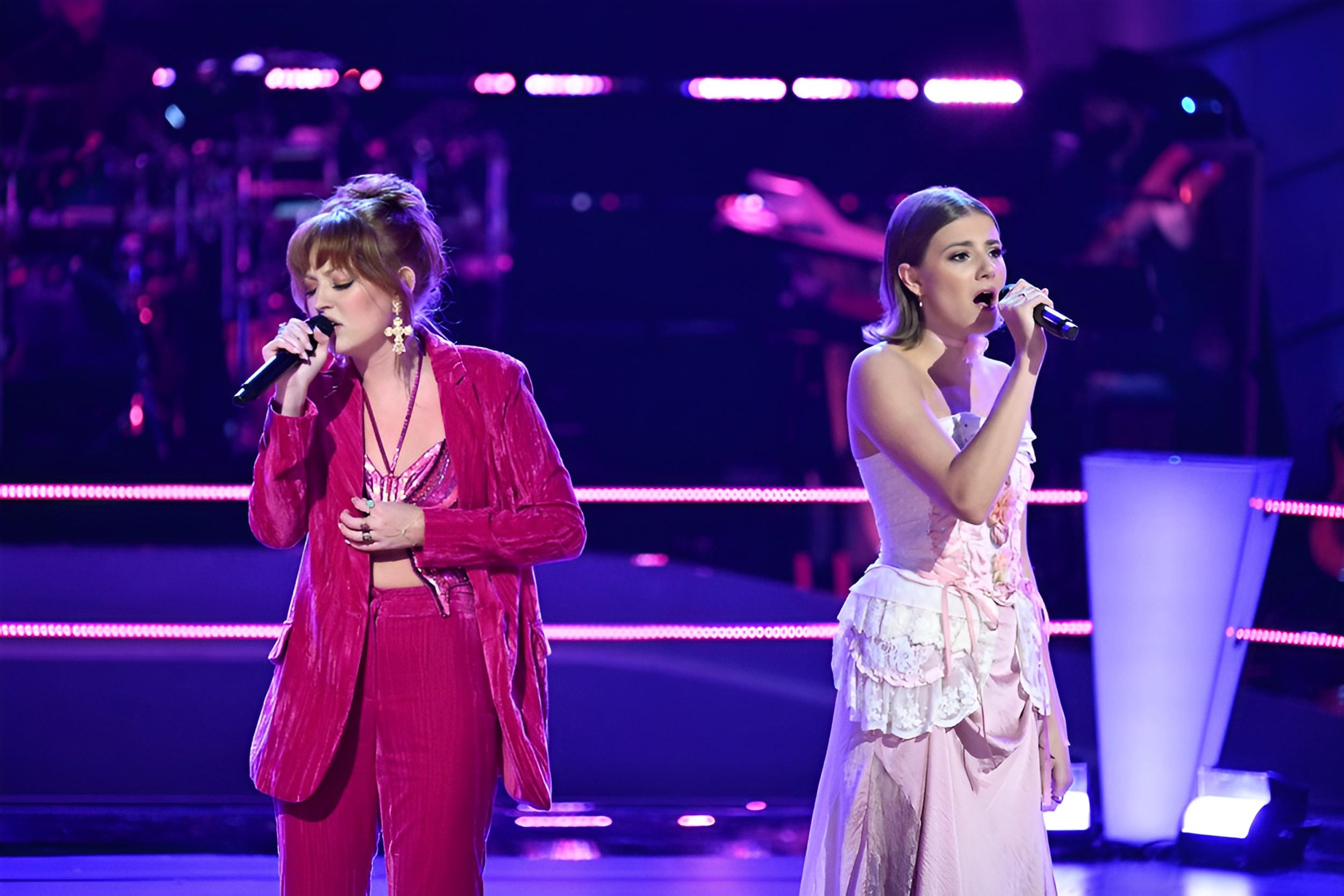 Iris Herrera, left, and Simone Marijic, right, stand on a stage lit by purple lights, each wearing pink and singing into a microphone.