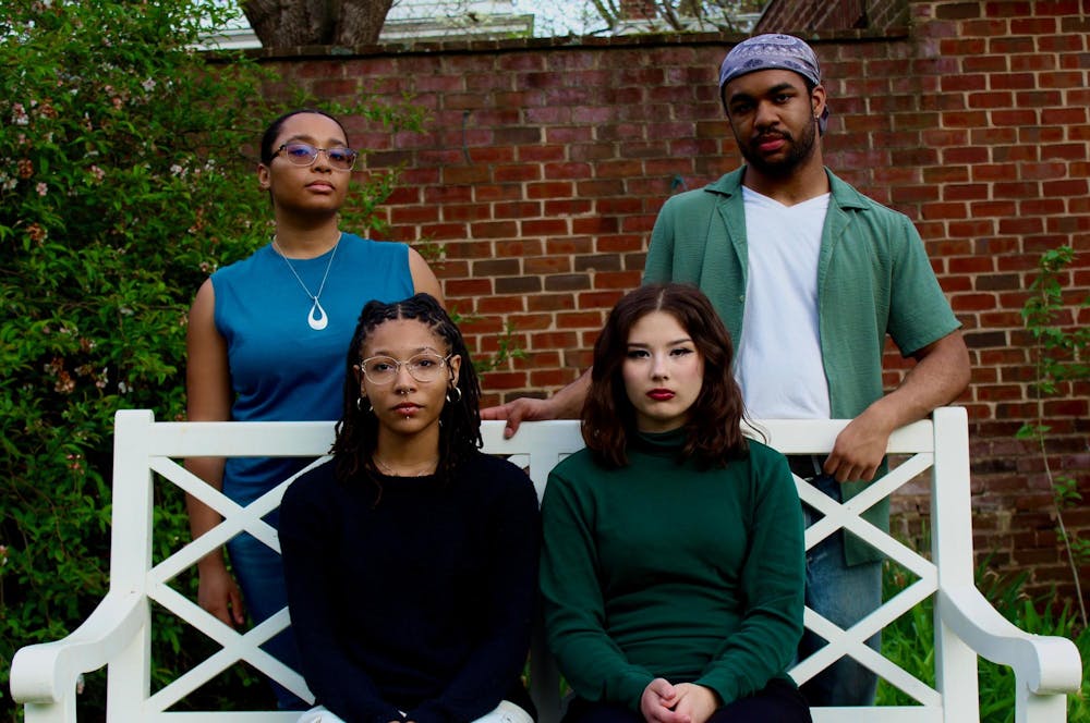 The four-person cast of "I Am My Mother's Daughter" pose in one of UVA's Gardens: two of them sit on a white bench, while two of them stand behind the bench.