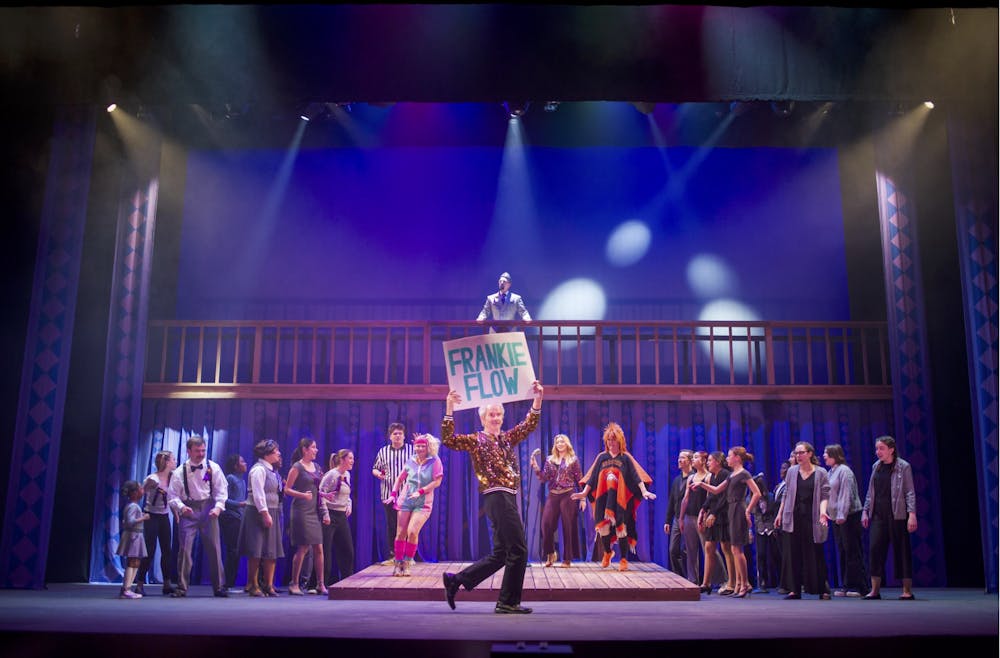 A crowd of performers stand on a stage. Above them, a man in a uniform looks down from a railing; in the center, a man holding a sign reading "FRANKIE FLOW" walks across the stage.