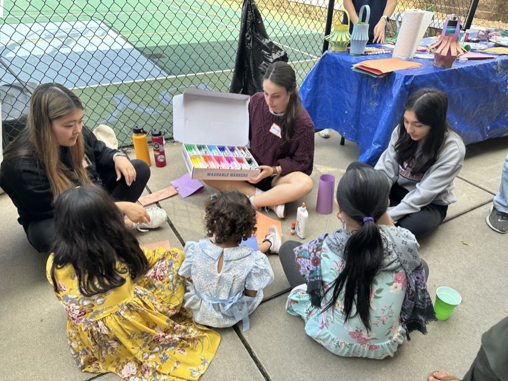 Volunteers from Cville Tulips sit outside with children, showing them a box of markers.