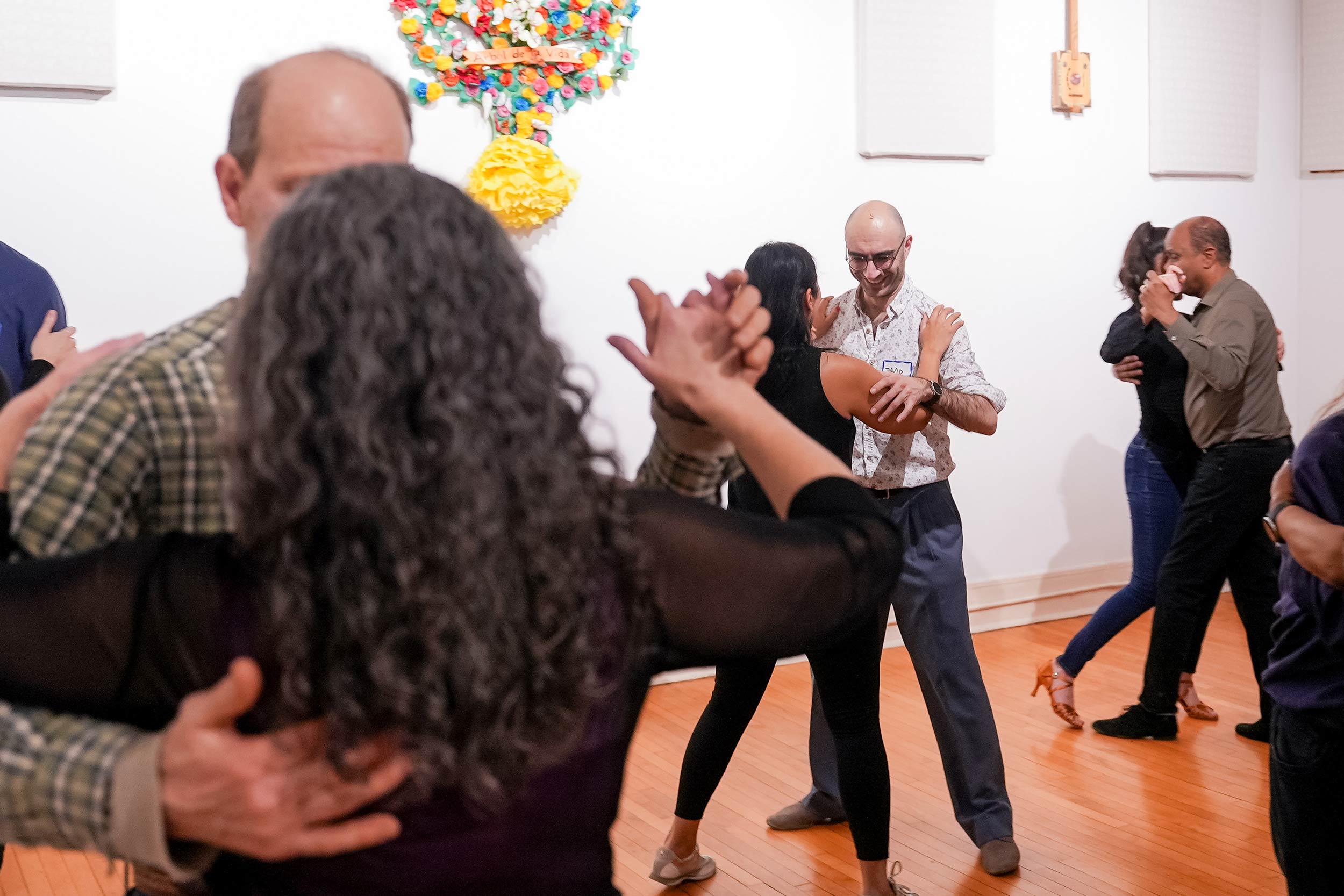 In a brightly lit room with white walls, many couples are learning to dance tango.