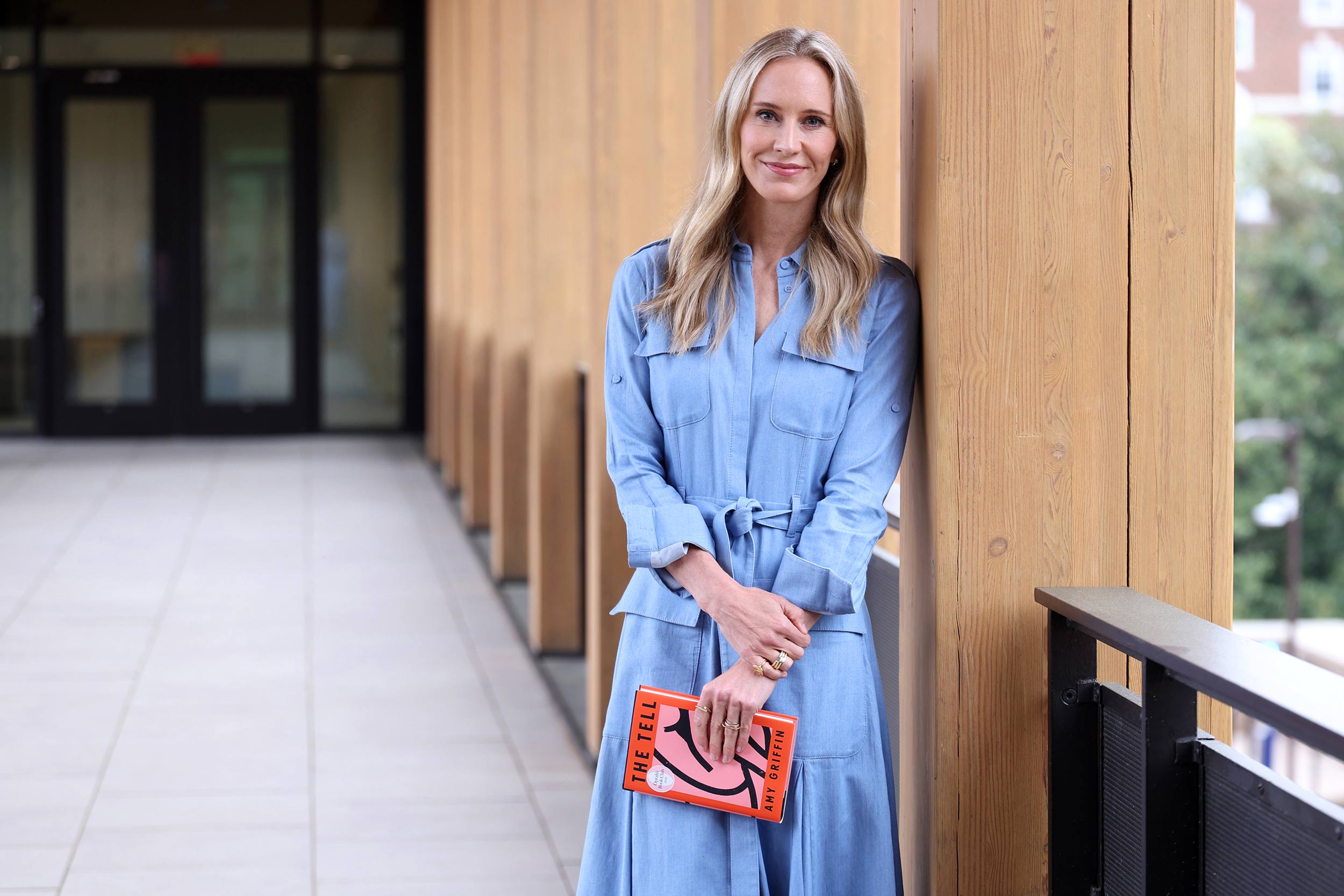 Amy Griffin leans against a wooden column while wearing a light blue dress and holding a copy of her book, The Tell.