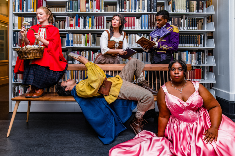 Five characters from Into the Woods are posing in front of a bookshelf in a library.