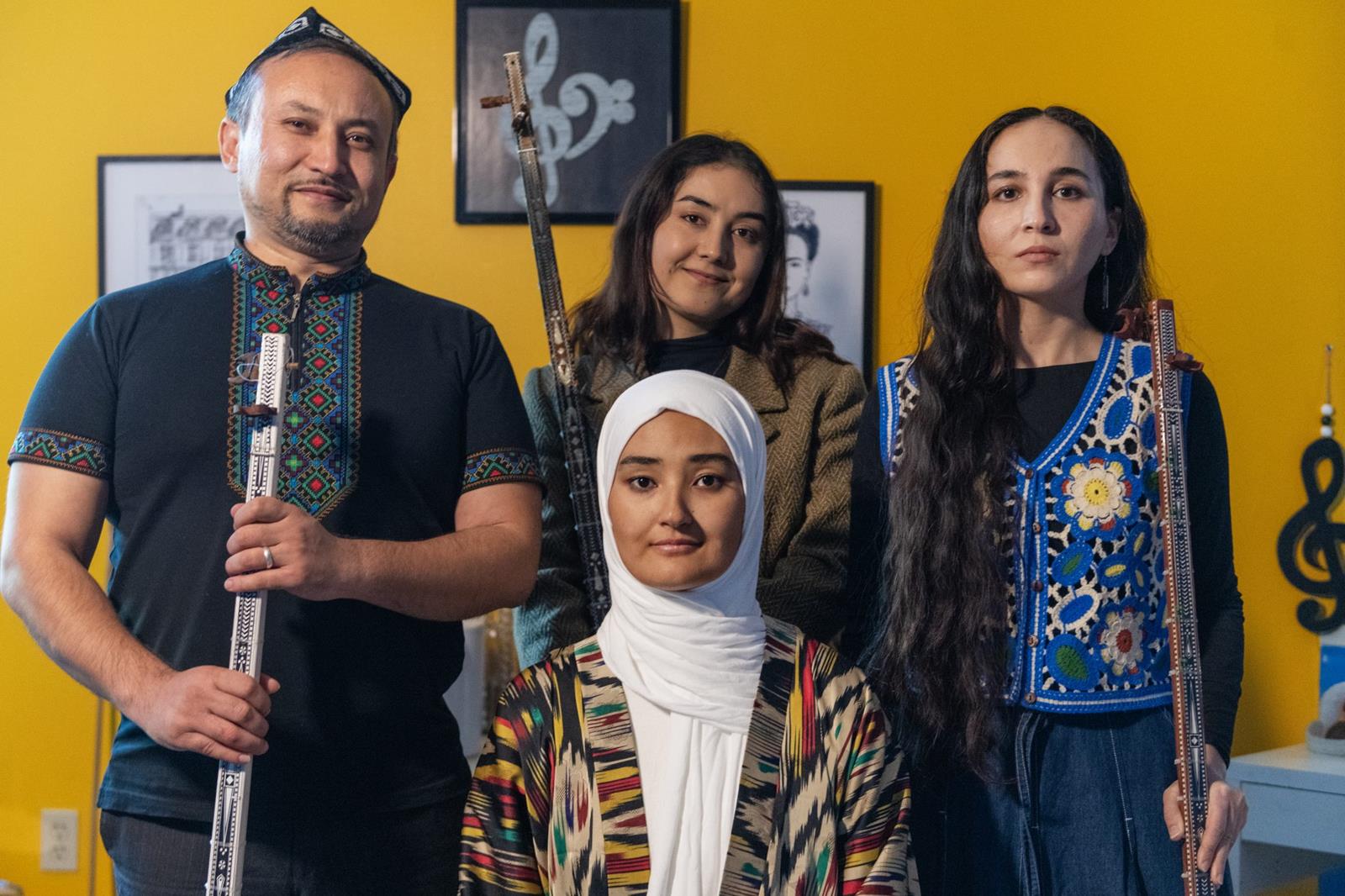 Hayrunisa Yenilmes, seated, and Amar Kilich, left, of Alexandria are apprenticing Ajinur Setiwaldi, right, and Subhinur Elemin of Arlington in Uyghur music traditions. The group is positioned in front of a bright yellow wall.