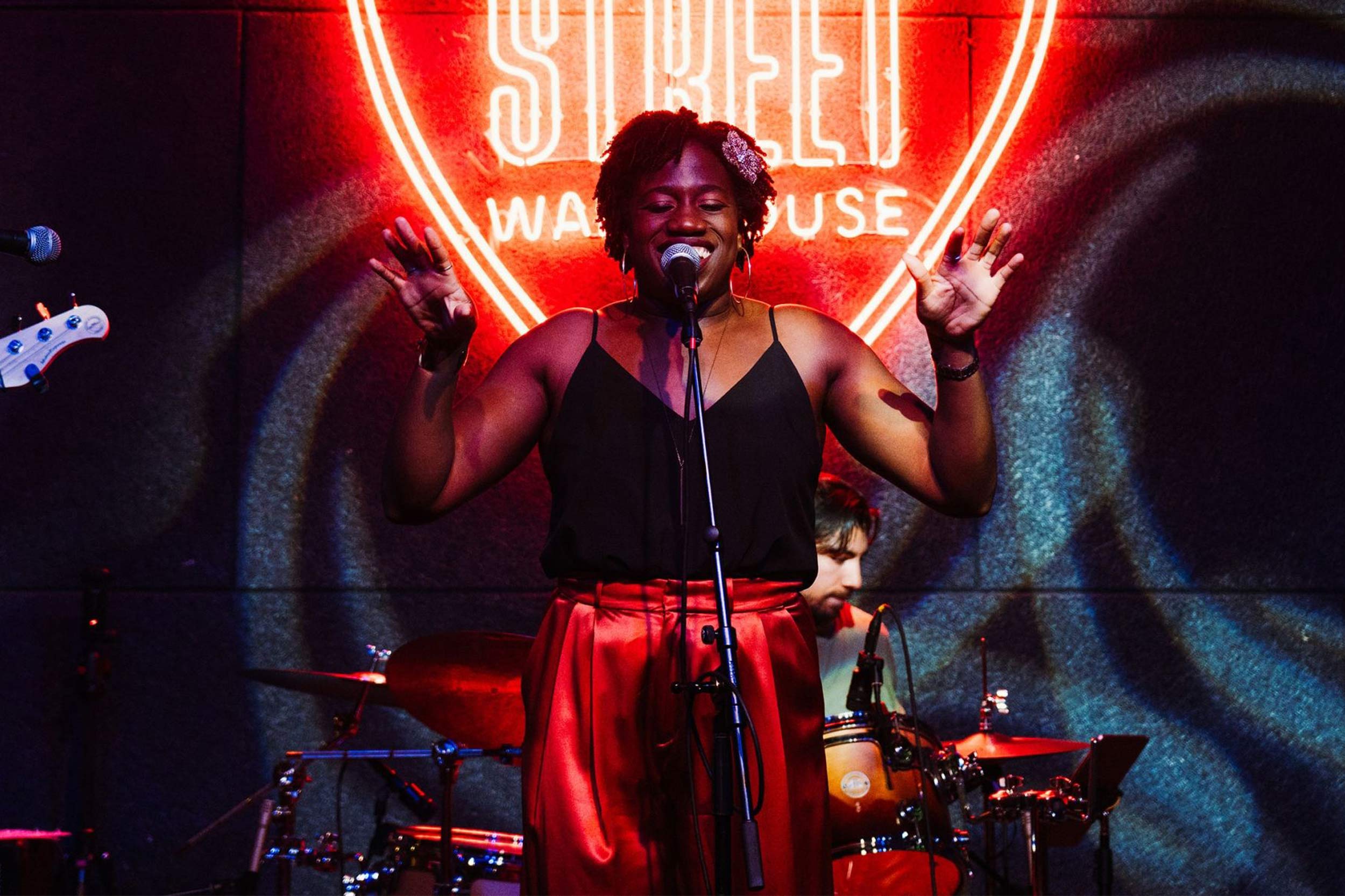Kemi Adegoroye stands at a microphone with both hands raised, standing in front of a drumset and a black wall with a bright neon-red letter sign.