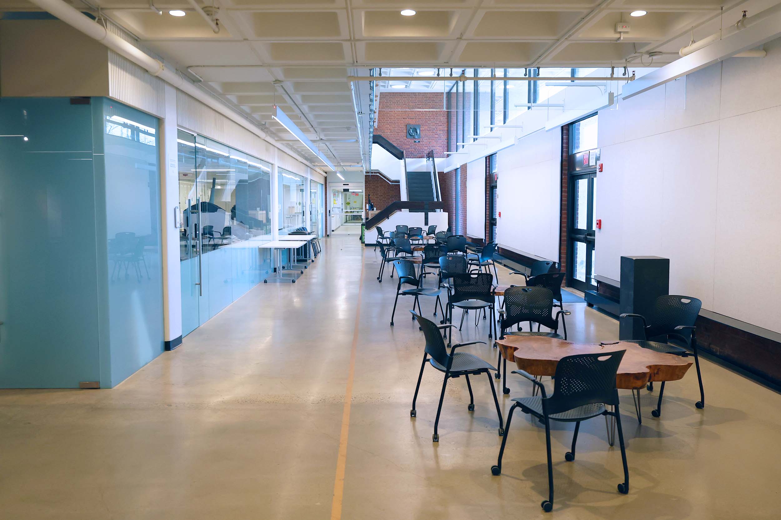 A lounge in the Architecture school has cement floors, various tables and chairs, a modern glass wall, and tall windows to let in ample sunlight.