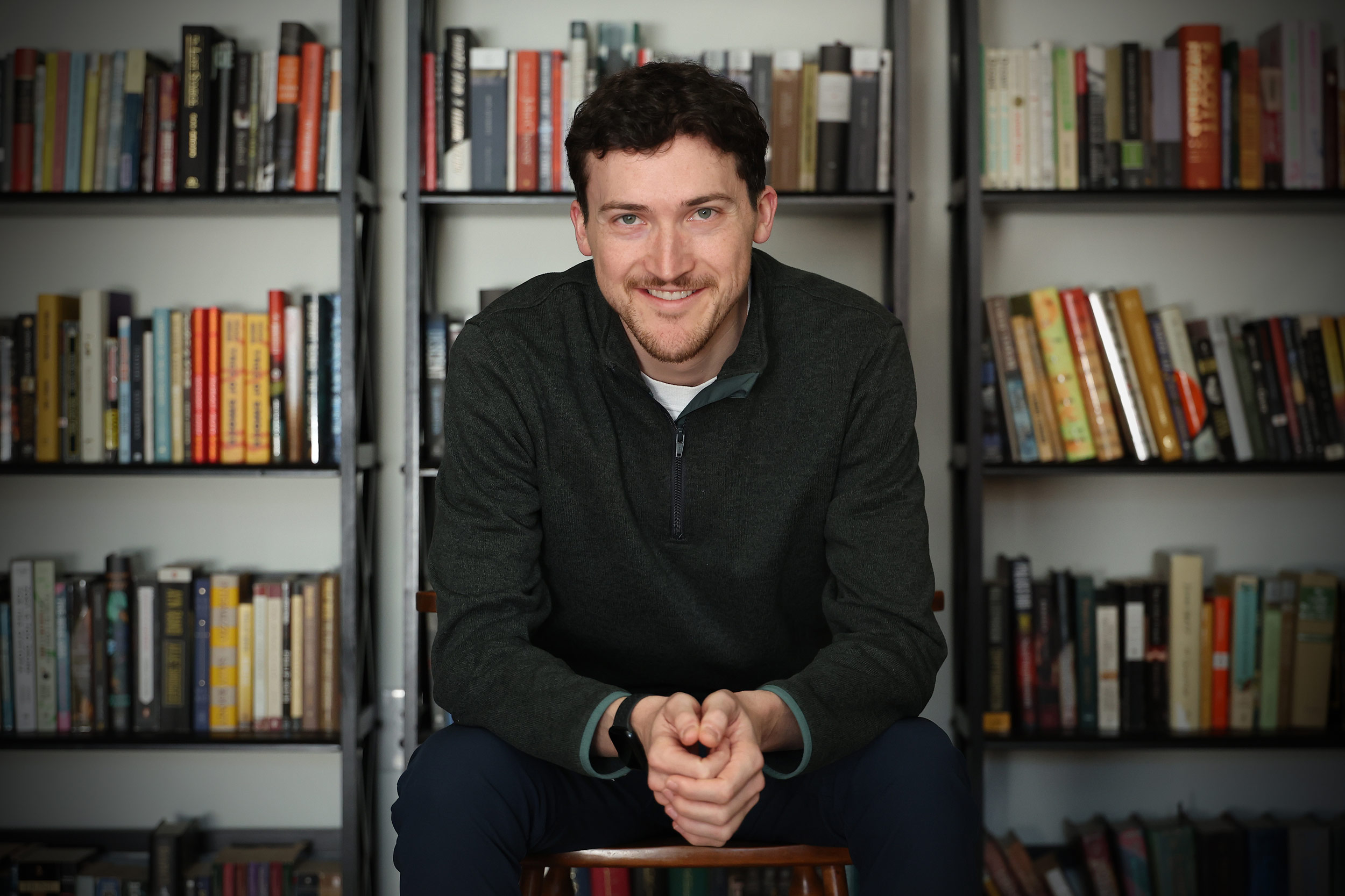 A photo of William Dozier sitting in front of a wall of bookshelves. He is leaning slightly forward with his hands clasped in front.