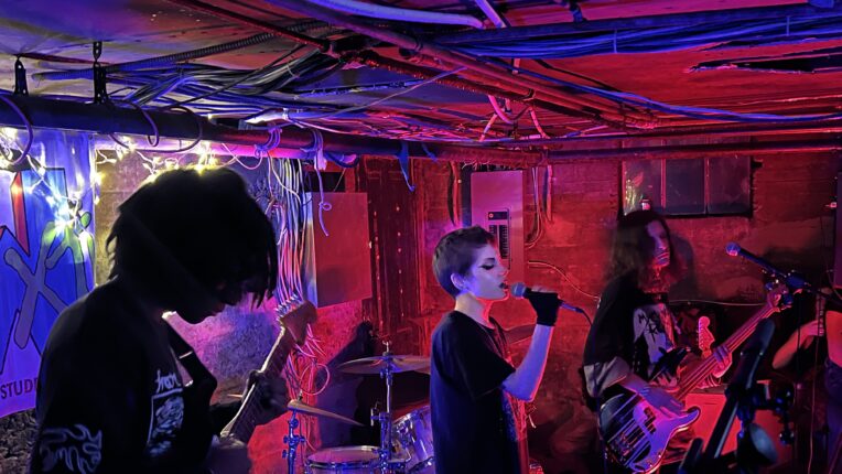 A basement show with band members performing, red and blue hues of light fill the space.
