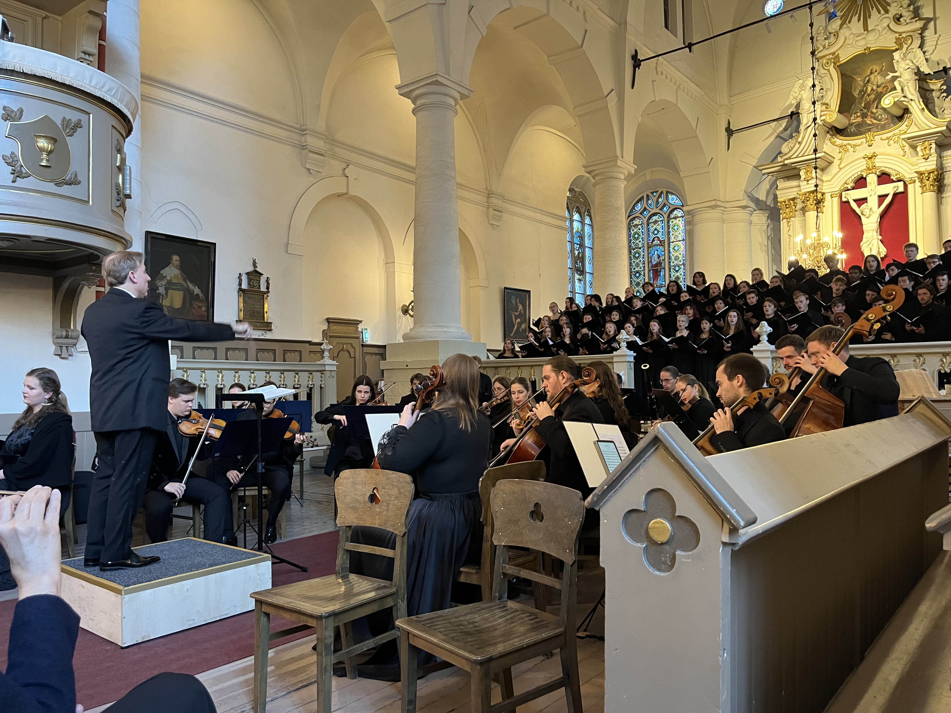Singers are shown standing in a church and performing, while members of an orchestra play instruments in front of them. The conductor is standing on an elevated platform.