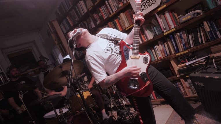A photo of a young man jamming on a red electric guitar while singing into a mic, positioned in front of a full bookshelf.