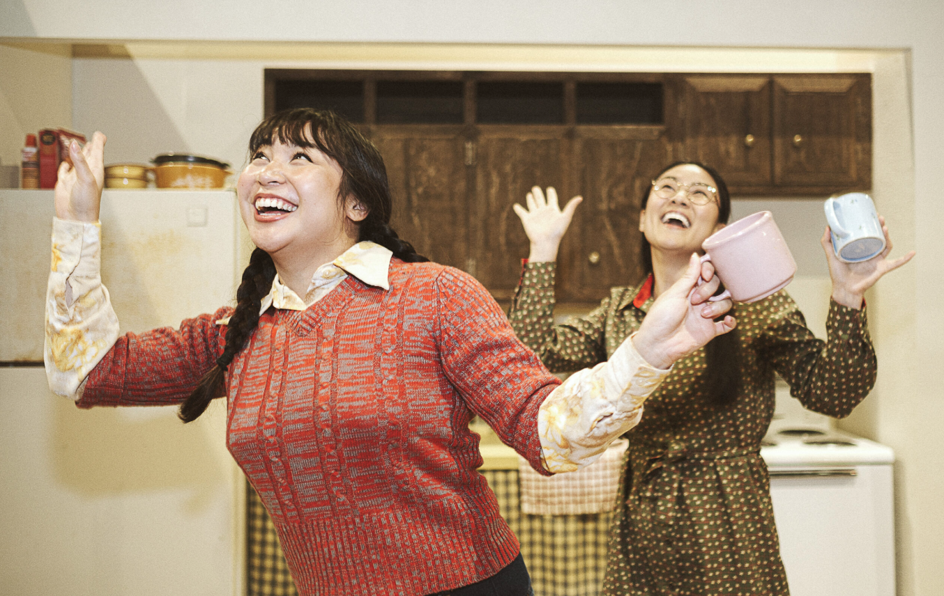 Two actresses from the show pose onstage, both holding coffee mugs in a kitchen, smiling and extending their arms.