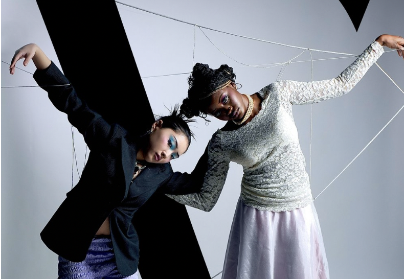 Two models are posed next to each other, leaning slightly down with their arms outstretched in a "broken doll" manner. 