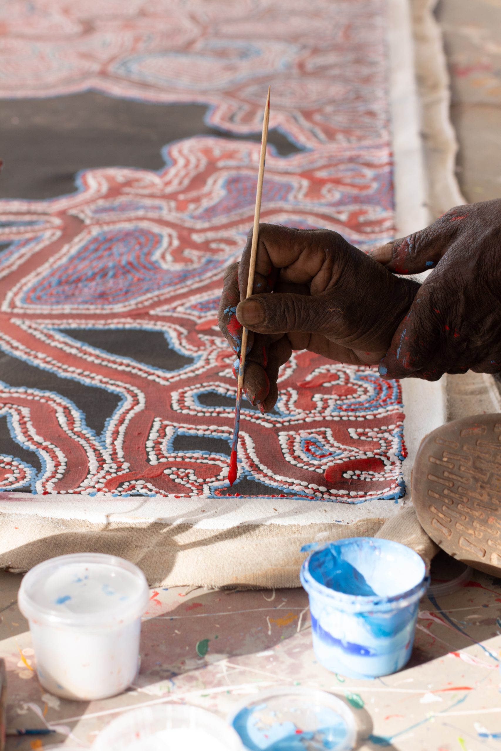 A close-up photo of a hand using a small paintbrush to paint a dotted design on a paper, using red, white, blue, and black.