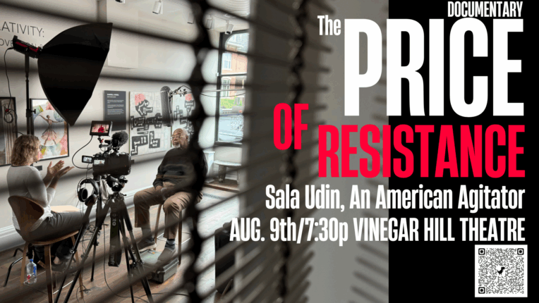 Photo taken through window blinds that show a film setup with a large light and camera, and one person interviewing another person while sitting.