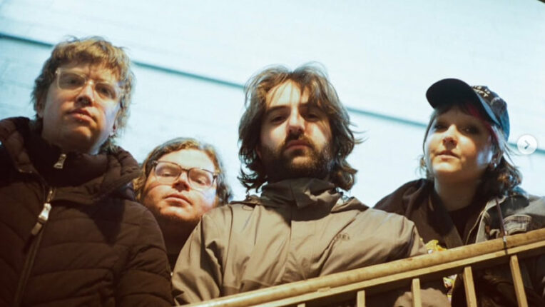 Four people, three men and one woman, are dressed in casual attire looking down into the camera lens with serious expressions.