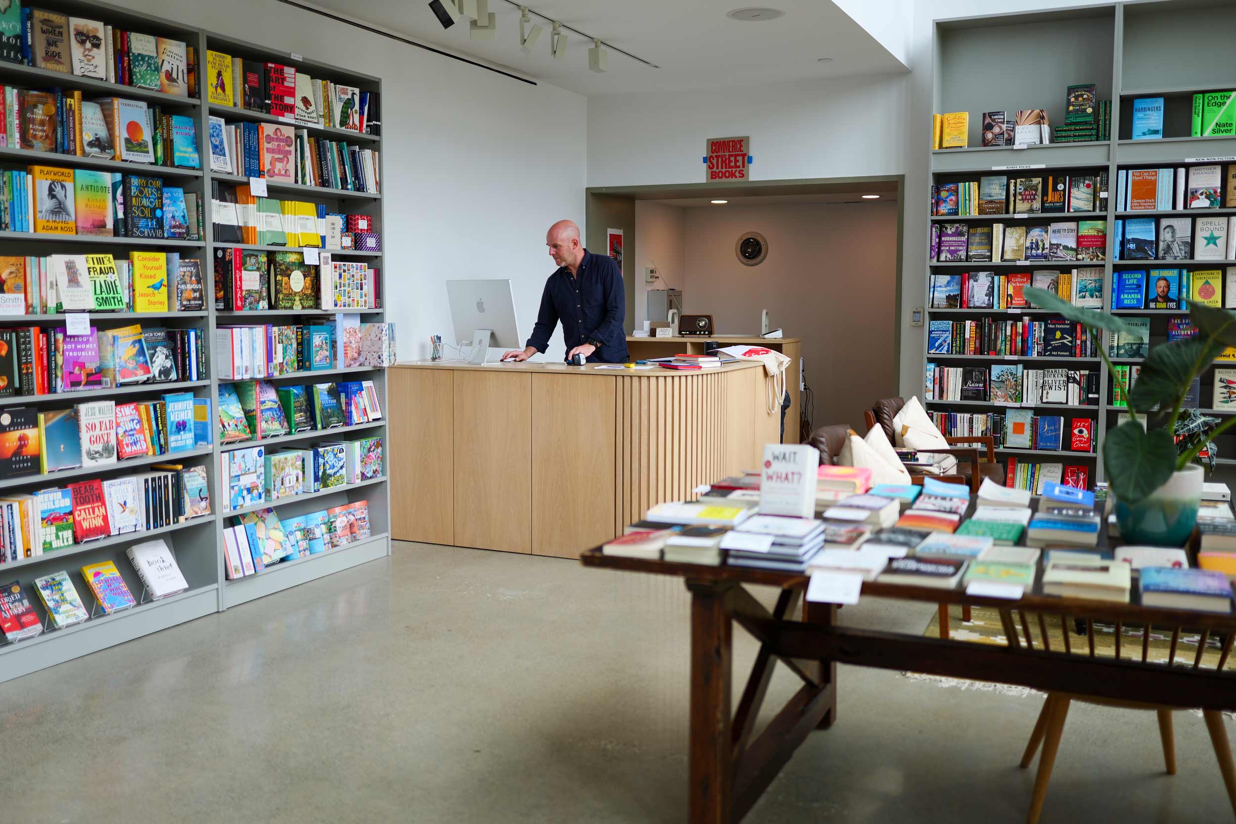 A room with white walls has large booksheleves filled with colorful books, and a table with more books displayed in the middle of the room. A man stands at the check-out counter and looks at a computer.