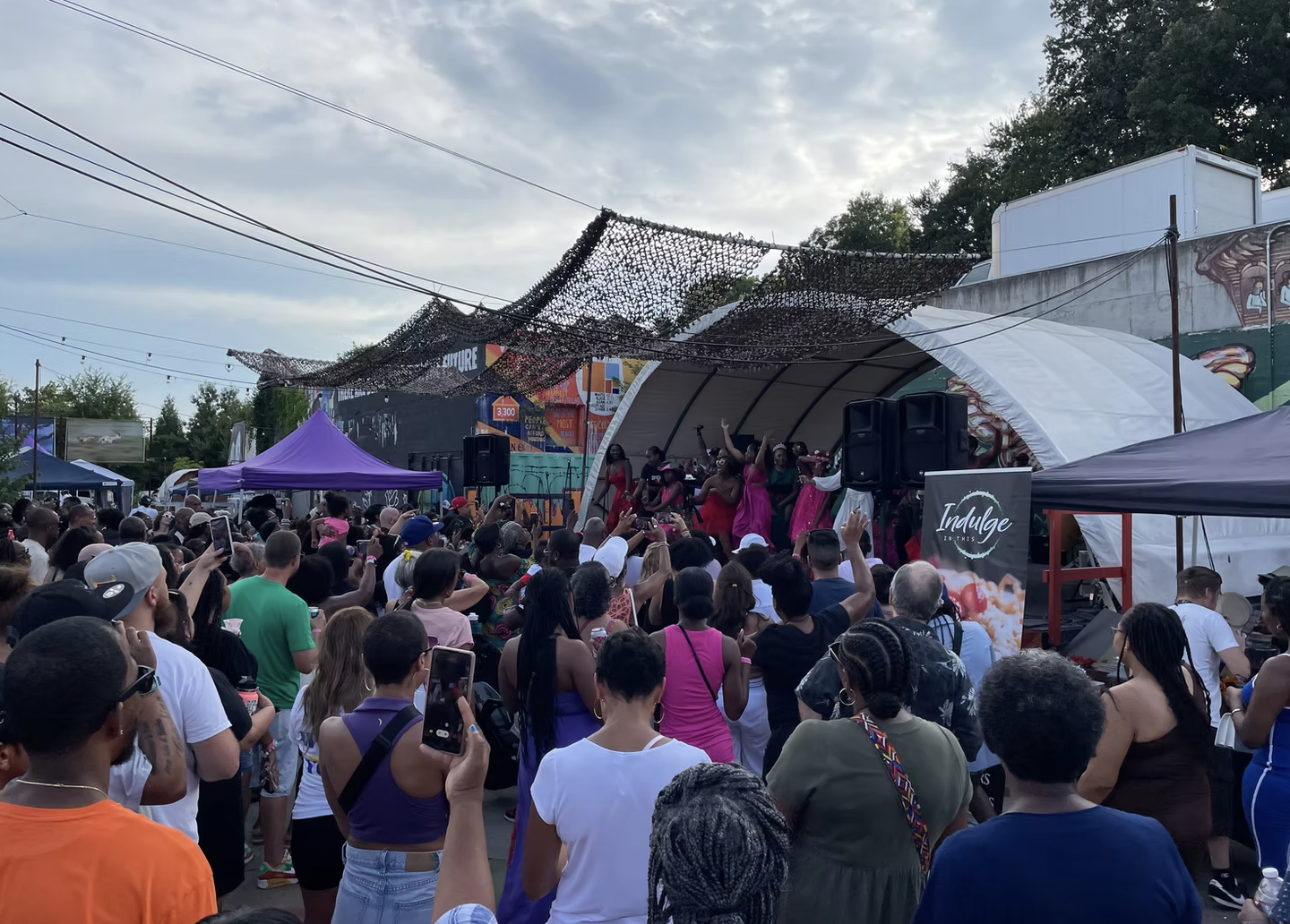A colorful crowd stands outside at IX Art Park in front of a covered stage.