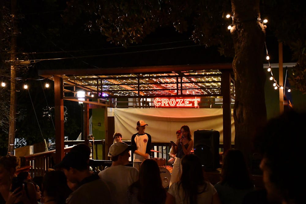 A picture of a small outdoor covered stage at night, with red neon sign at the back that says "CROZET" and has 3 people talking on stage.