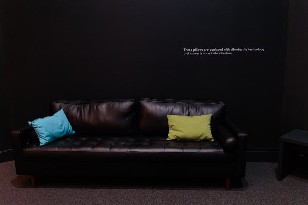 A very dark room with only a black leather couch. Two small rectangular pillows are on the couch, one green and one blue.