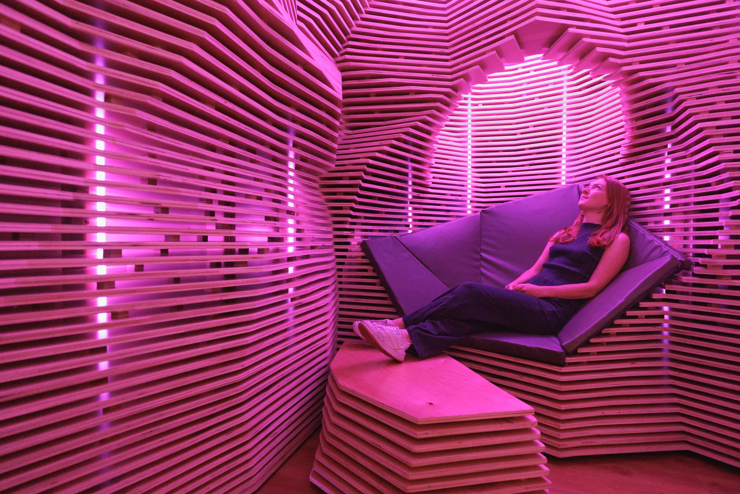 A room with textured wooden-slat walls has a padded nook in the wall for sitting. The lights are a purple/pink hue.