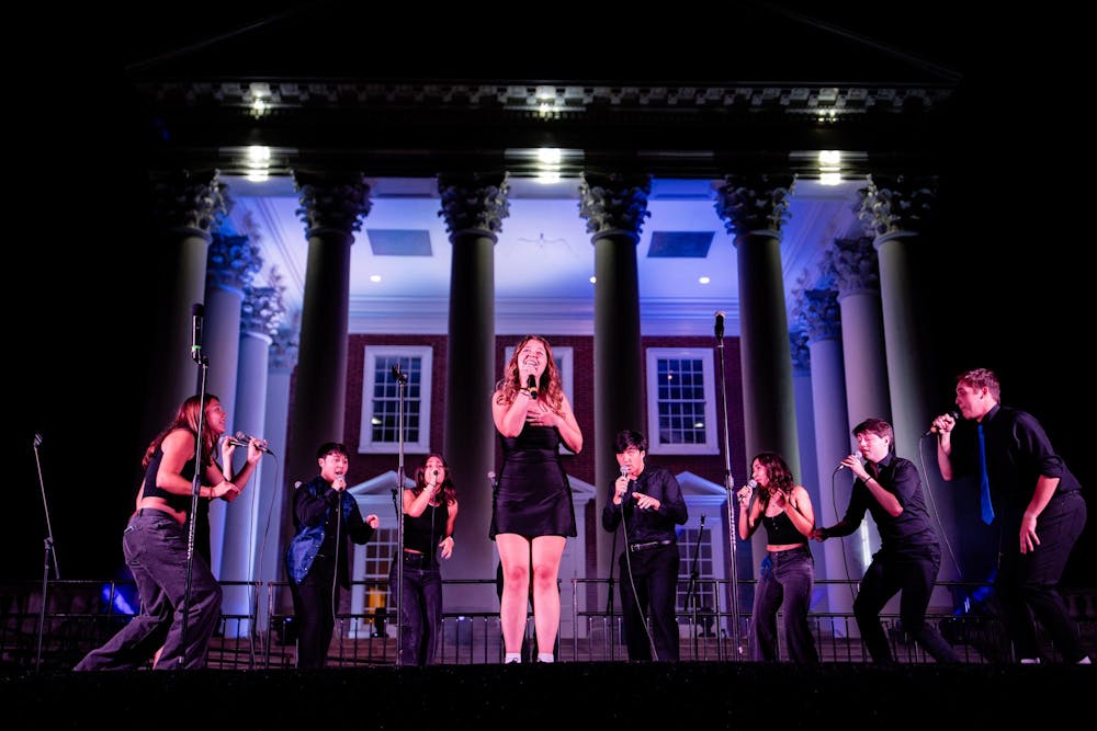 A group of students standing in a semi-circle sing on stage in front of the Rotunda. There are blue hues of light on the students and Rotunda.