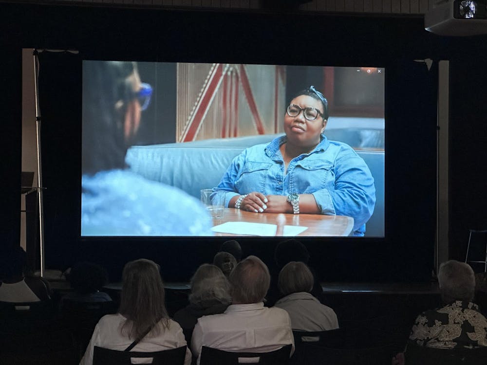 A movie screen with a woman sitting at a restaurant booth, and we can see the audience watching the screen from a viewpoint of the back row.