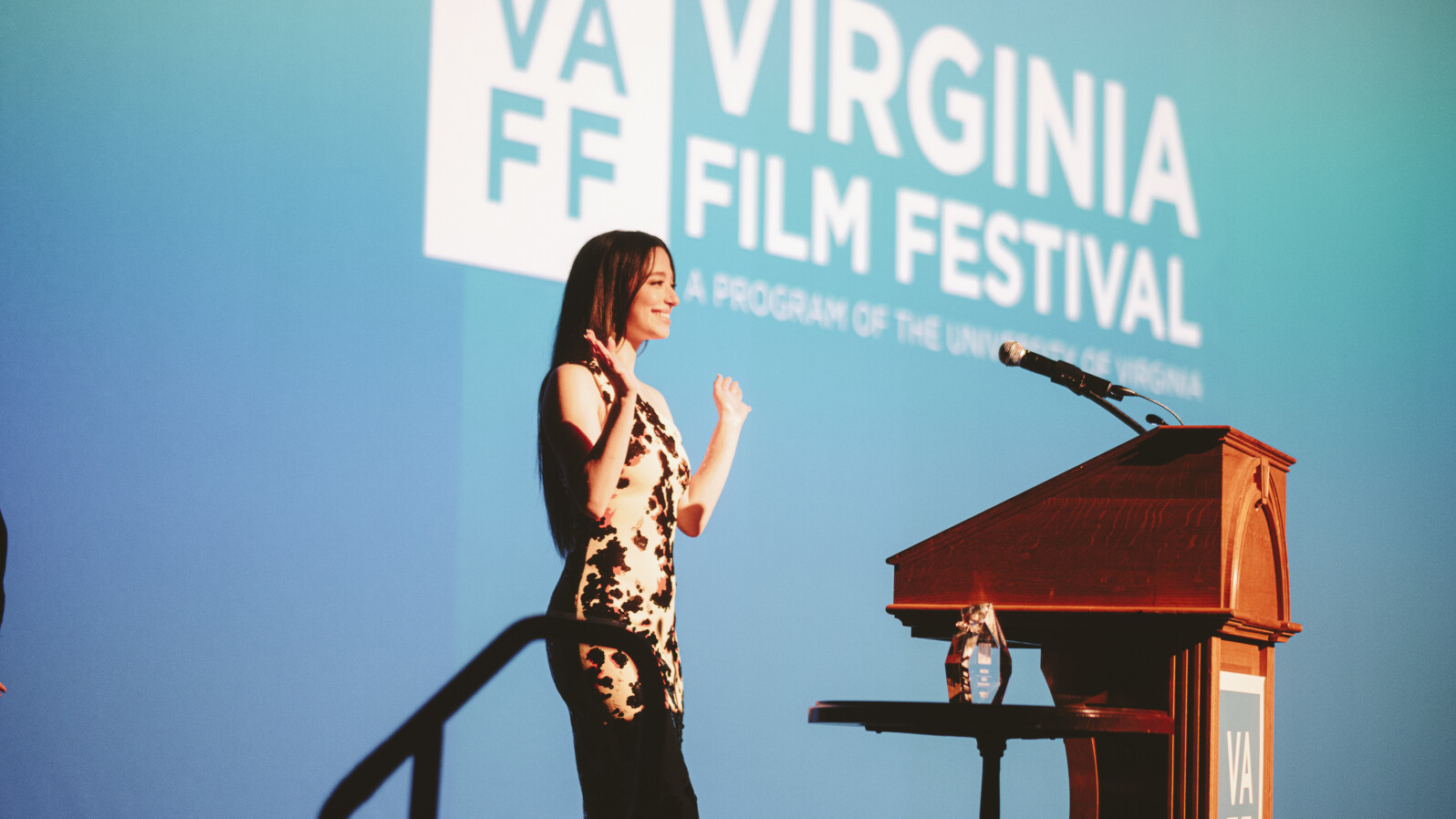A woman walks up to a podium waving to a audience out of view, with the text "Virginia Film Festival" projected on a blue background behind her.