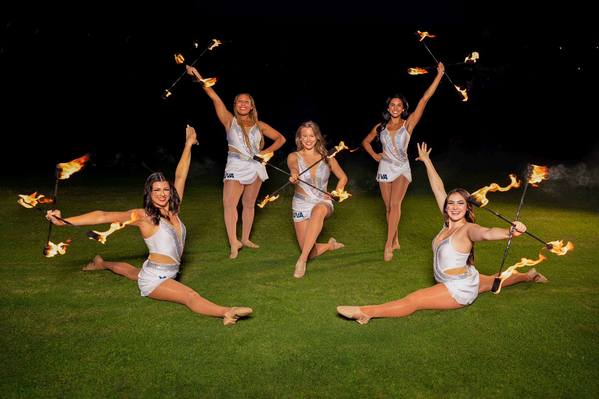 Five young women in dance leotards are posed on a football field holding flaming batons. 