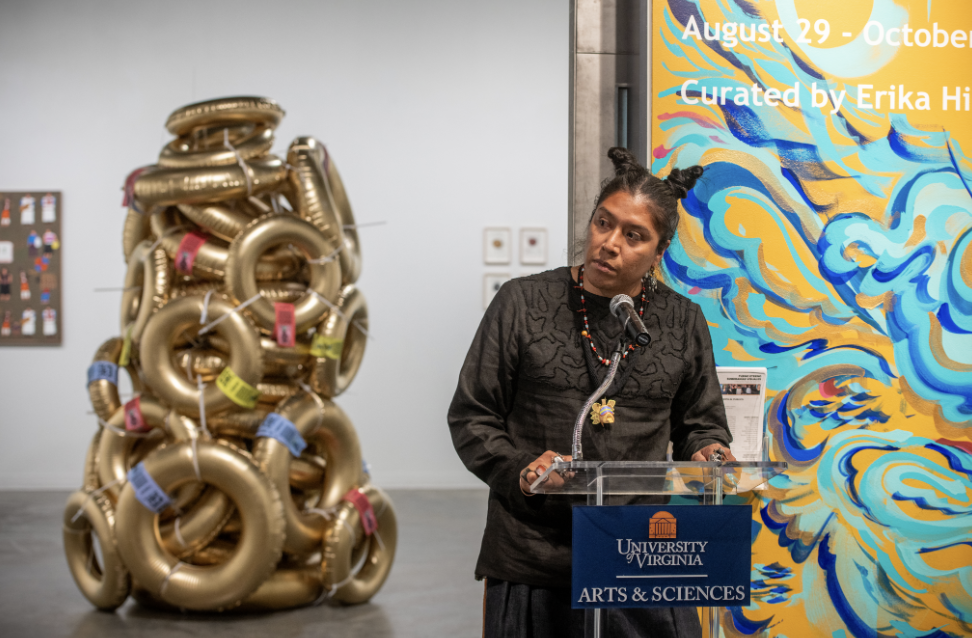 A woman stands at a podium speaking at an art exhibition, with a tall pile of golden inner-tubes stacked in the gallery behind her.