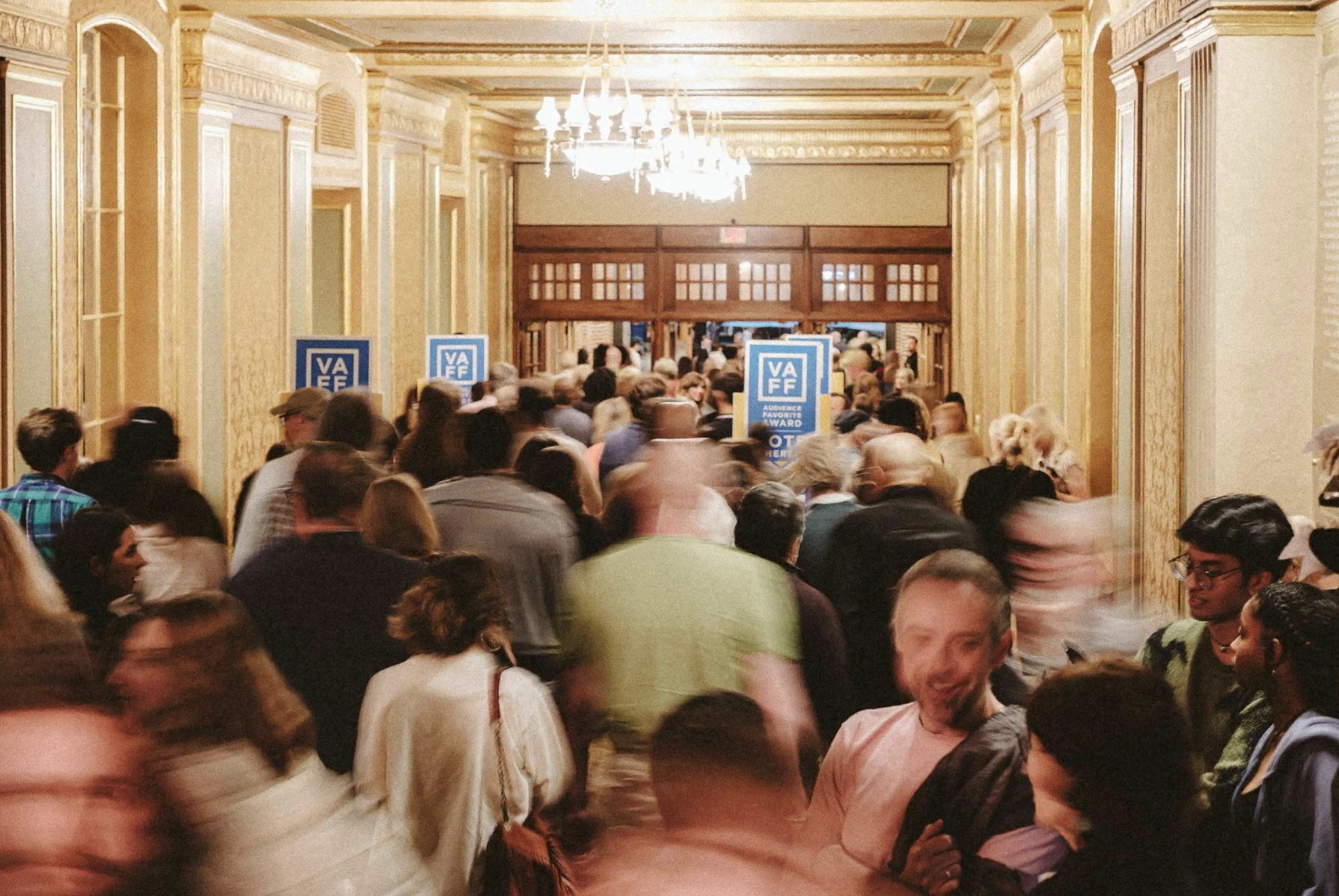 The hallway of a nice theatre venue that is filled with people rushing about, with chandeliers hanging from the ceiling, and various blue signs for the Virginia Film Festival.