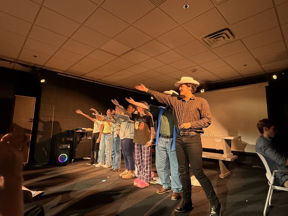 A small black box theatre shows a line of performers at bows, pointing to the back of the theatre to recognize their director. The lights are directed at the performers.