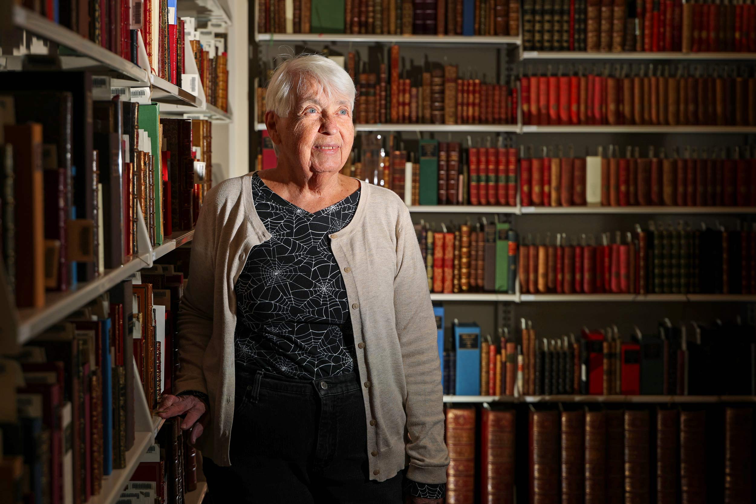 An older woman with short white hair stands amongst bookshelves and looks off to the side.