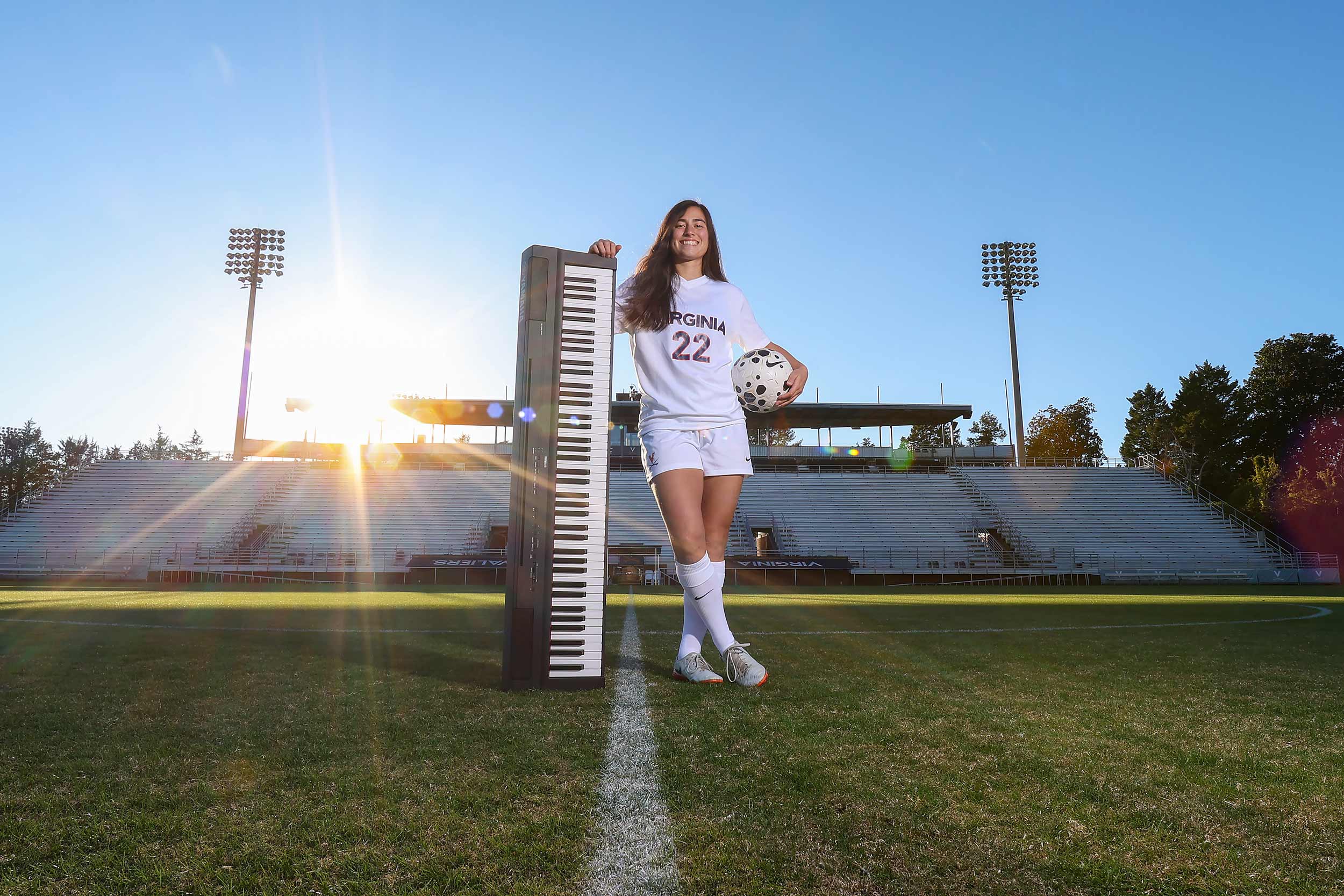 A UVA female soccer player with long brown hair stands on the field and is hold a soccer ball under one arm, and is leaning on a propped-up keyboard under the other arm.