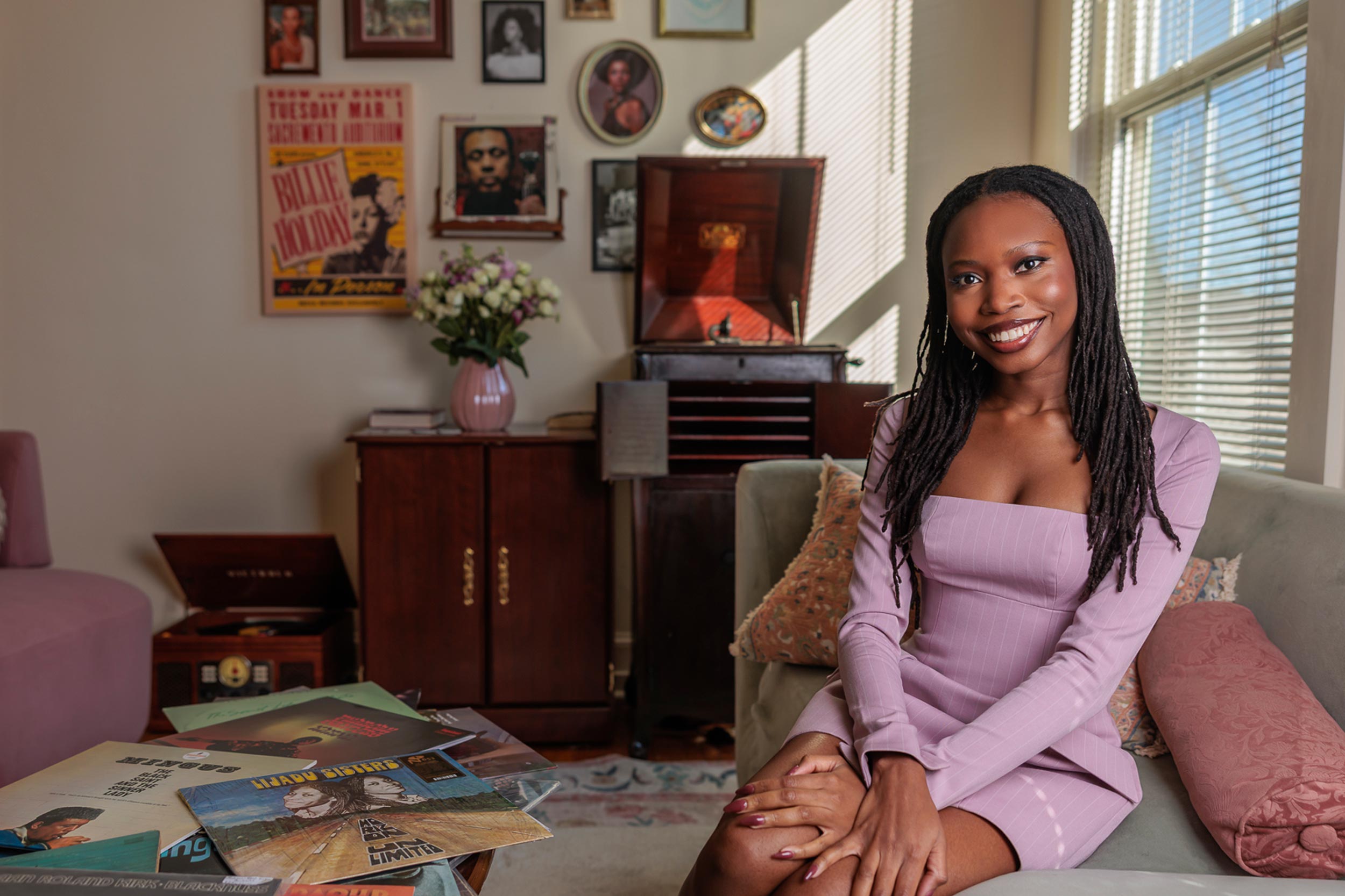 The interior of a girl's bedroom where she sits smiling on a small couch, and behind her we see a record player with various posters on the wall and vinyls spread out on the coffee table.